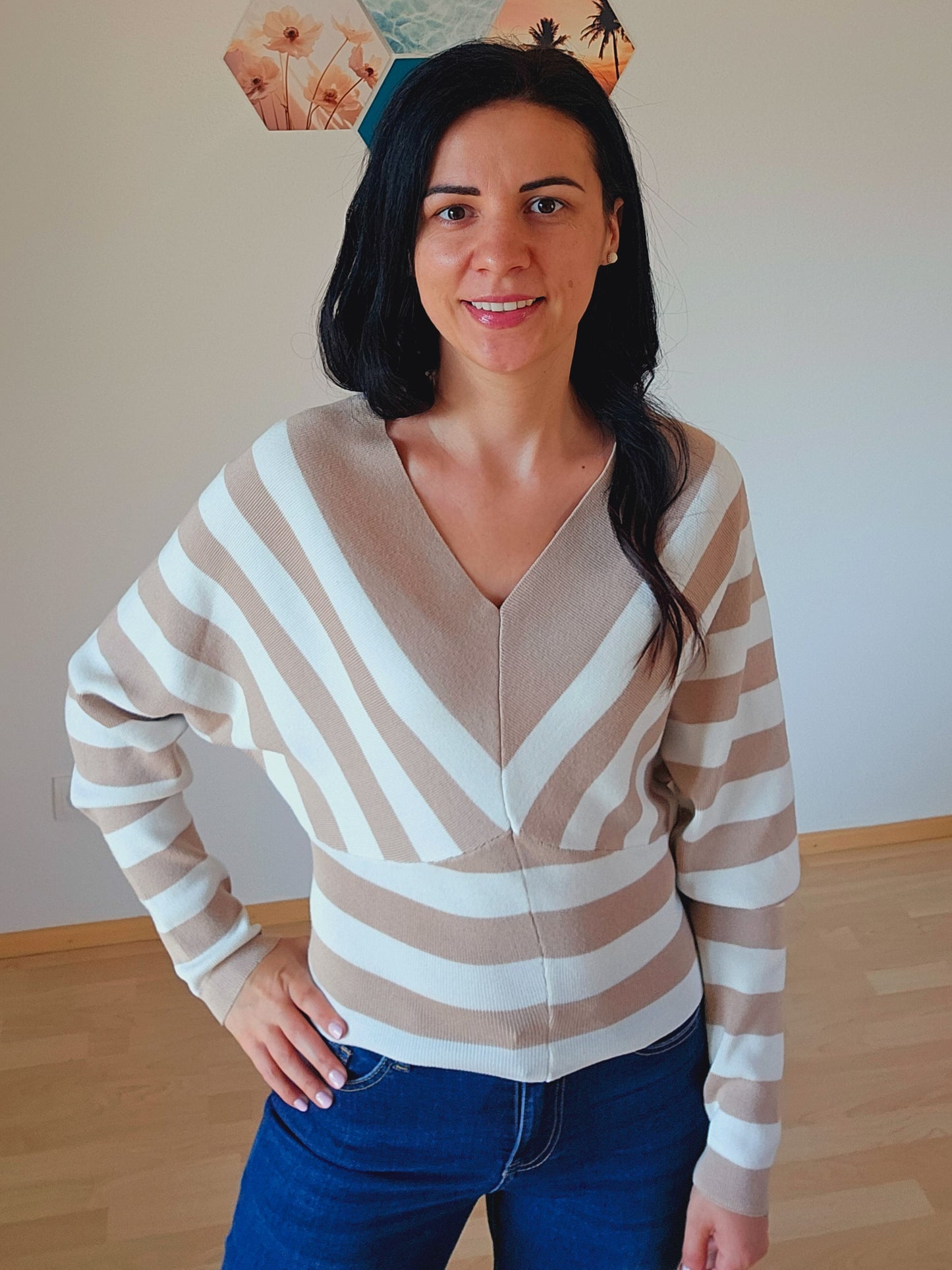 Das Model trägt einen gestreiften Pullover mit V-Ausschnitt in Beige und Weiß. Die breiten Streifen verlaufen diagonal und horizontal, was dem Oberteil ein interessantes Design verleiht.