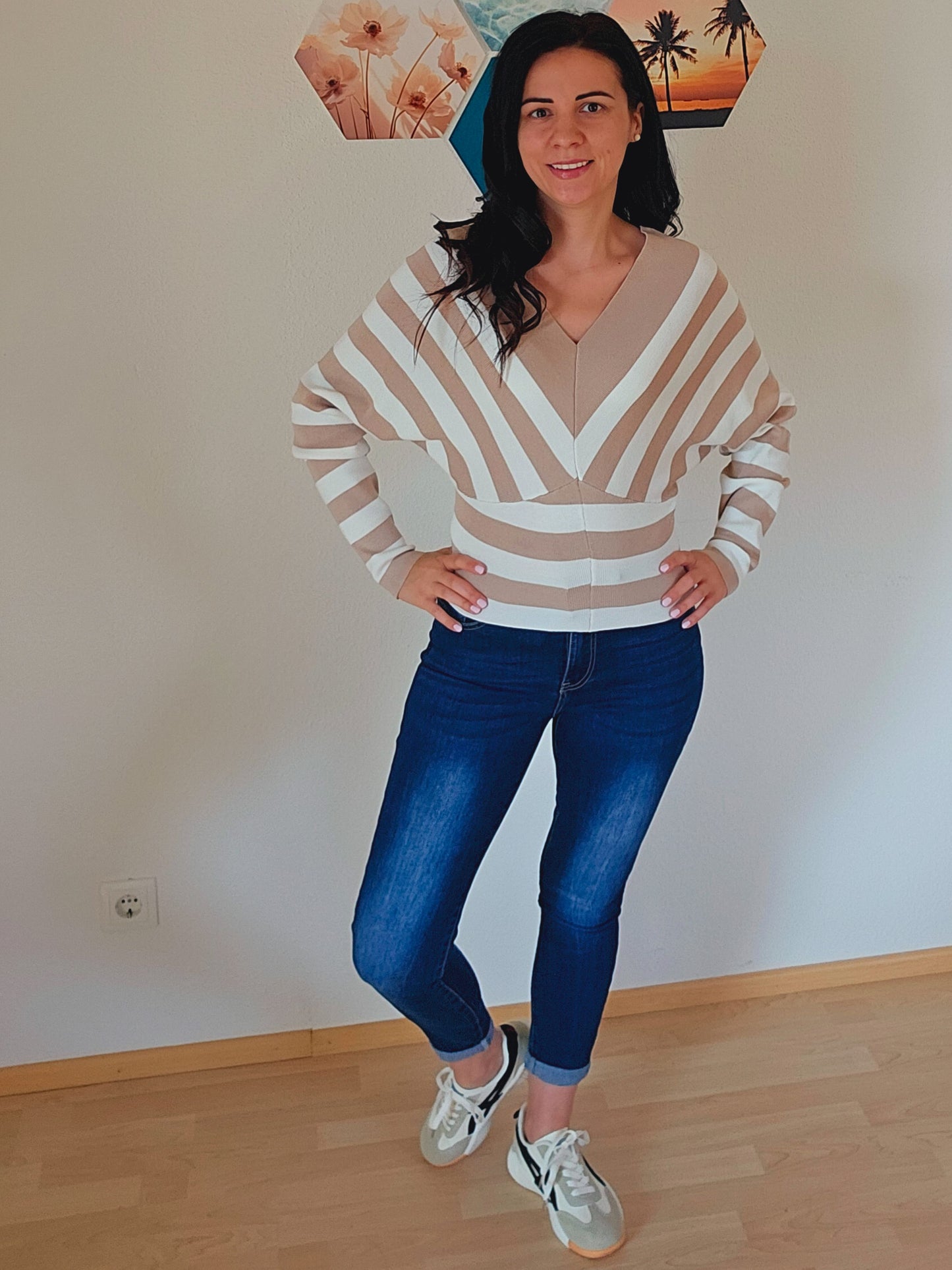 Das Model trägt einen gestreiften Pullover mit V-Ausschnitt in Beige und Weiß. Die breiten Streifen verlaufen diagonal und horizontal, was dem Oberteil ein interessantes Design verleiht.
