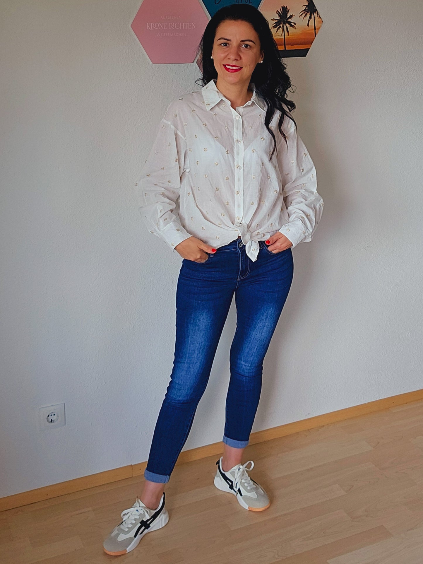 Das Model trägt eine weiße Bluse mit dezenten golden Punkten. Die Bluse ist geknotet und hat einen klassischen Kragen und Knopfleiste. Dazu trägt die Person eine dunkelblaue, schmal geschnittene Jeans. Der Look ist lässig und elegant.