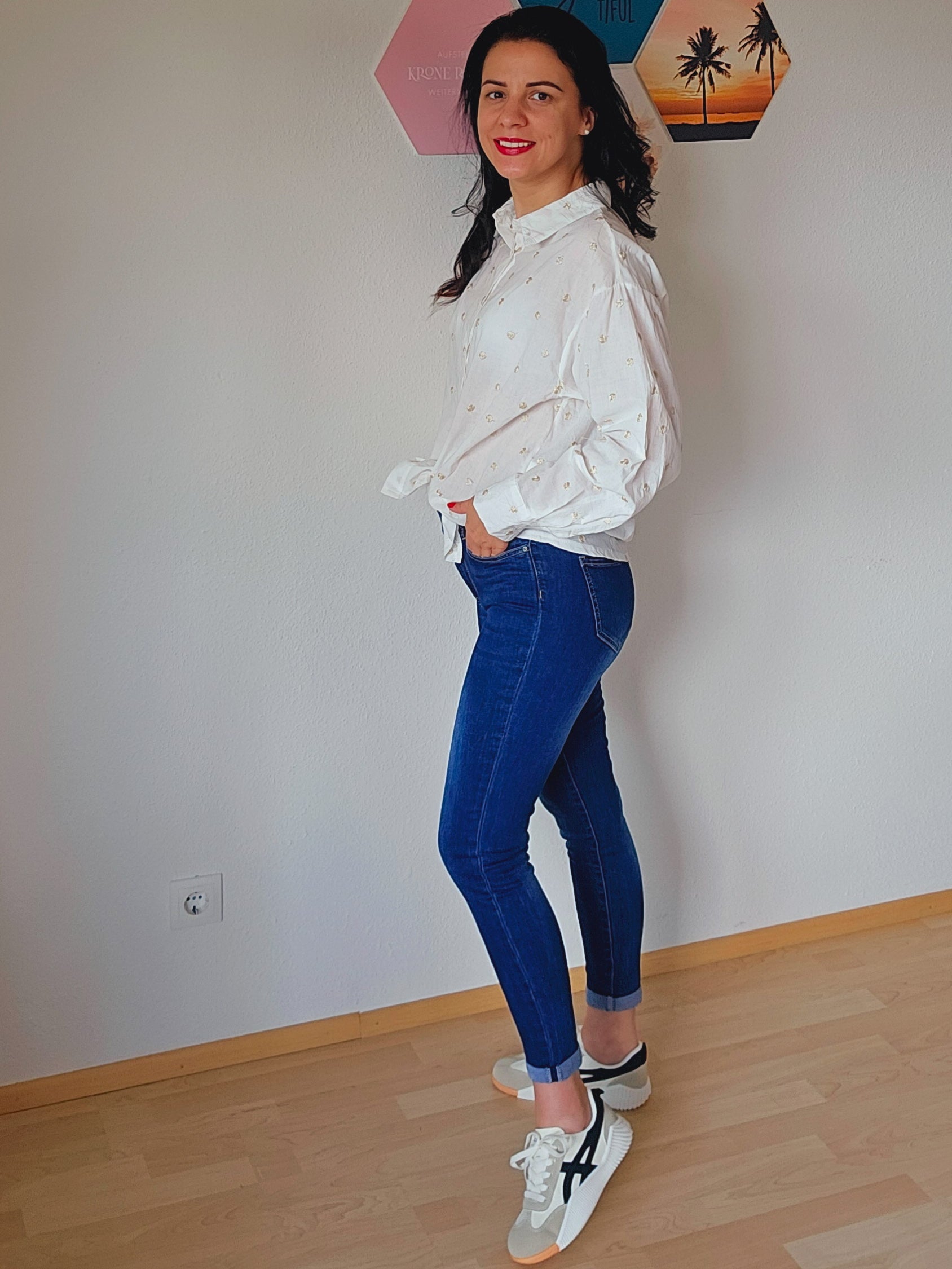 Das Model trägt eine weiße Bluse mit dezenten golden Punkten. Die Bluse ist geknotet und hat einen klassischen Kragen und Knopfleiste. Dazu trägt die Person eine dunkelblaue, schmal geschnittene Jeans. Der Look ist lässig und elegant.