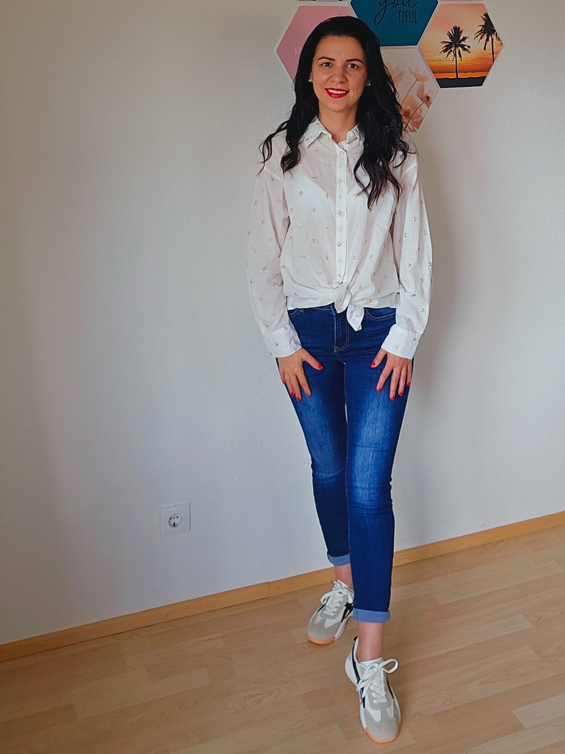 Das Model trägt eine weiße Bluse mit dezenten golden Punkten. Die Bluse ist geknotet und hat einen klassischen Kragen und Knopfleiste. Dazu trägt die Person eine dunkelblaue, schmal geschnittene Jeans. Der Look ist lässig und elegant.