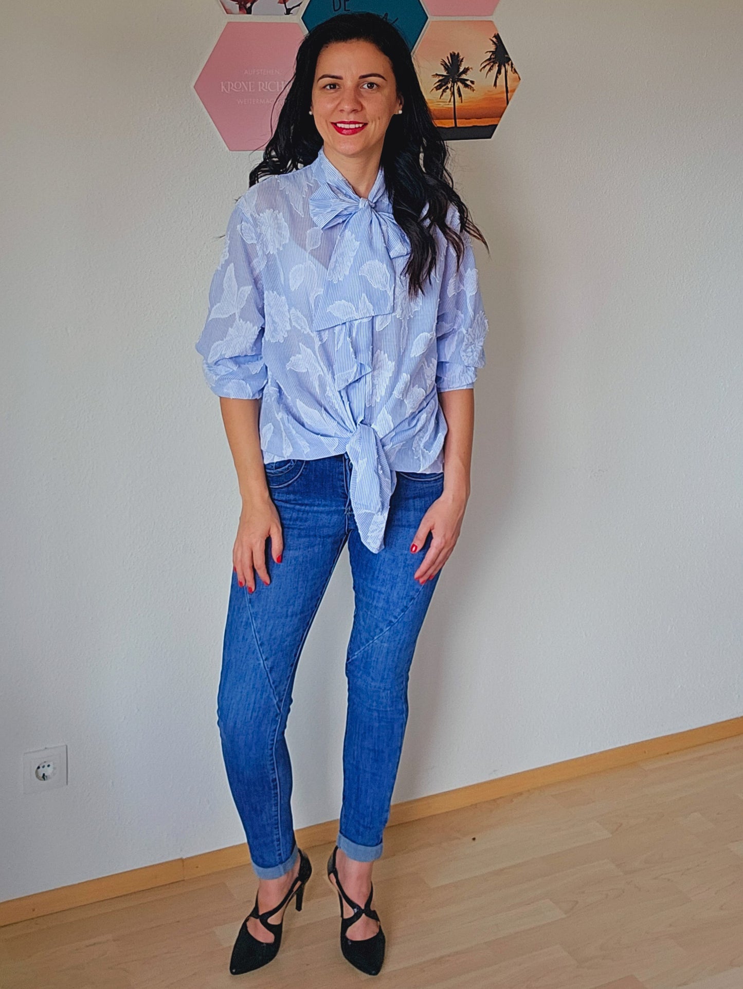 Das Model trägt eine hellblaue Bluse mit einem floralen Muster und einer auffälligen Schleife am Kragen. Sie ist lässig geknotet. Dazu trägt die Person eine blaue, schmal geschnittene Jeans und schwarze, elegante Pumps. Der Look ist eine stilvolle Kombination aus casual und schick.