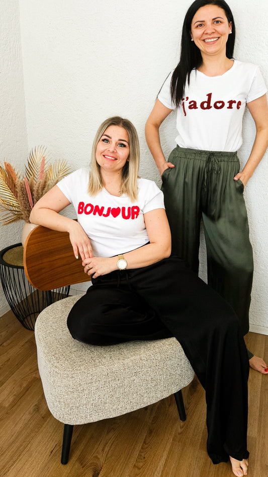 Auf dem Bild sind 2 Frauen zu sehen. Eine sitzt auf einem Sessel und trägt ein basic weißes T-Shirt mit dem Aufdruck 'Bonjour' in roter Farbe und eine legere schwarze satin Hose. Die andere steht hinter ihr und trägt ein basic weißes T-Shirt mit dem Aufdruck 'j´adore' in wineroter Farbe und eine legere  olive satin Hose.